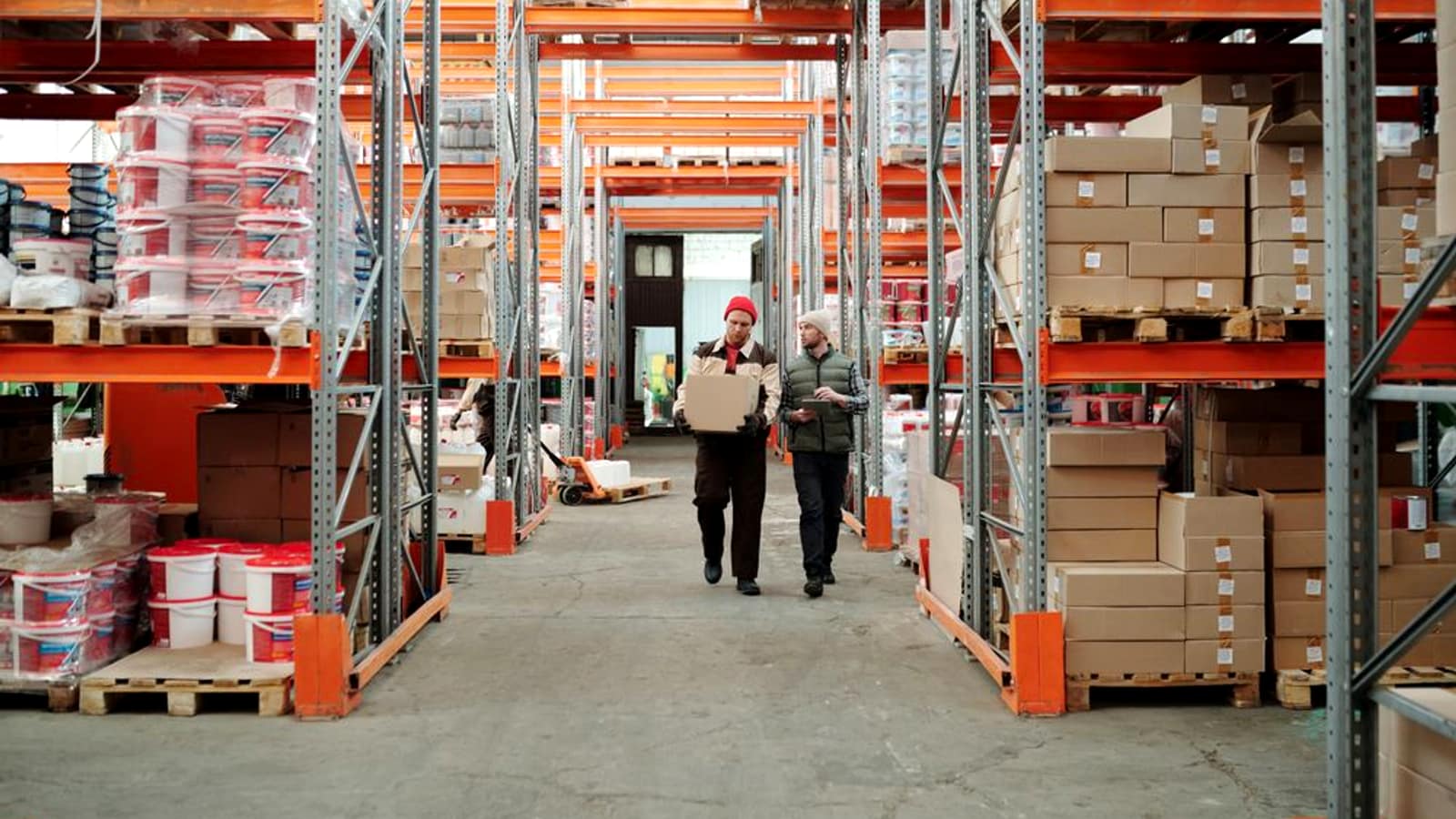 Two workers walking down an aisle of a warehouse stocked with pallets and boxes on tall racks