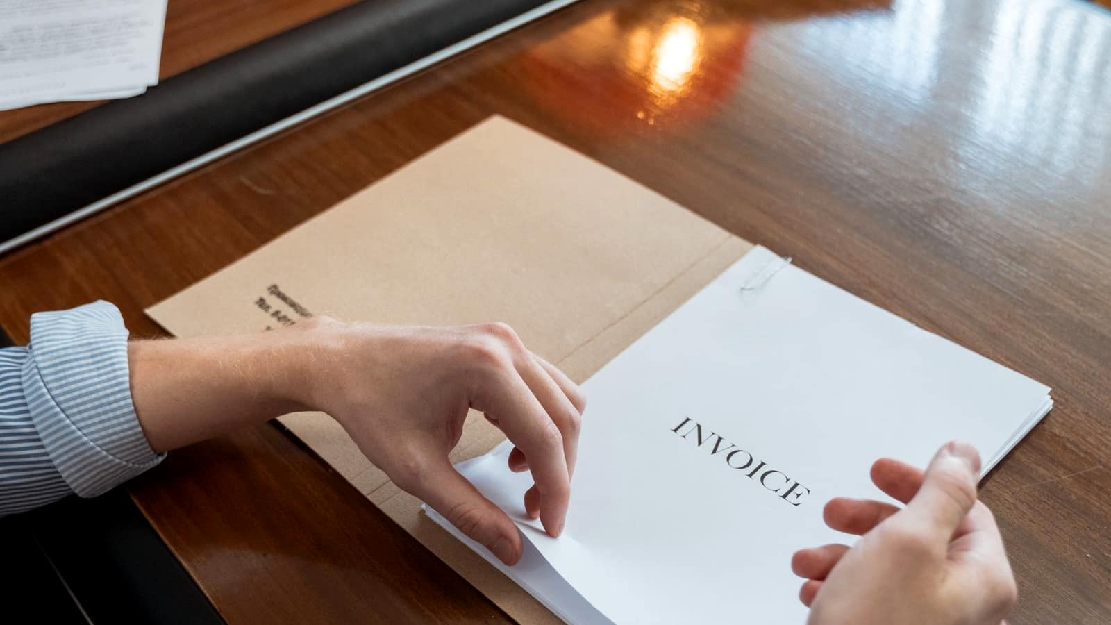 A person's hands resting on a printed document headed INVOICE on a dark wood desk