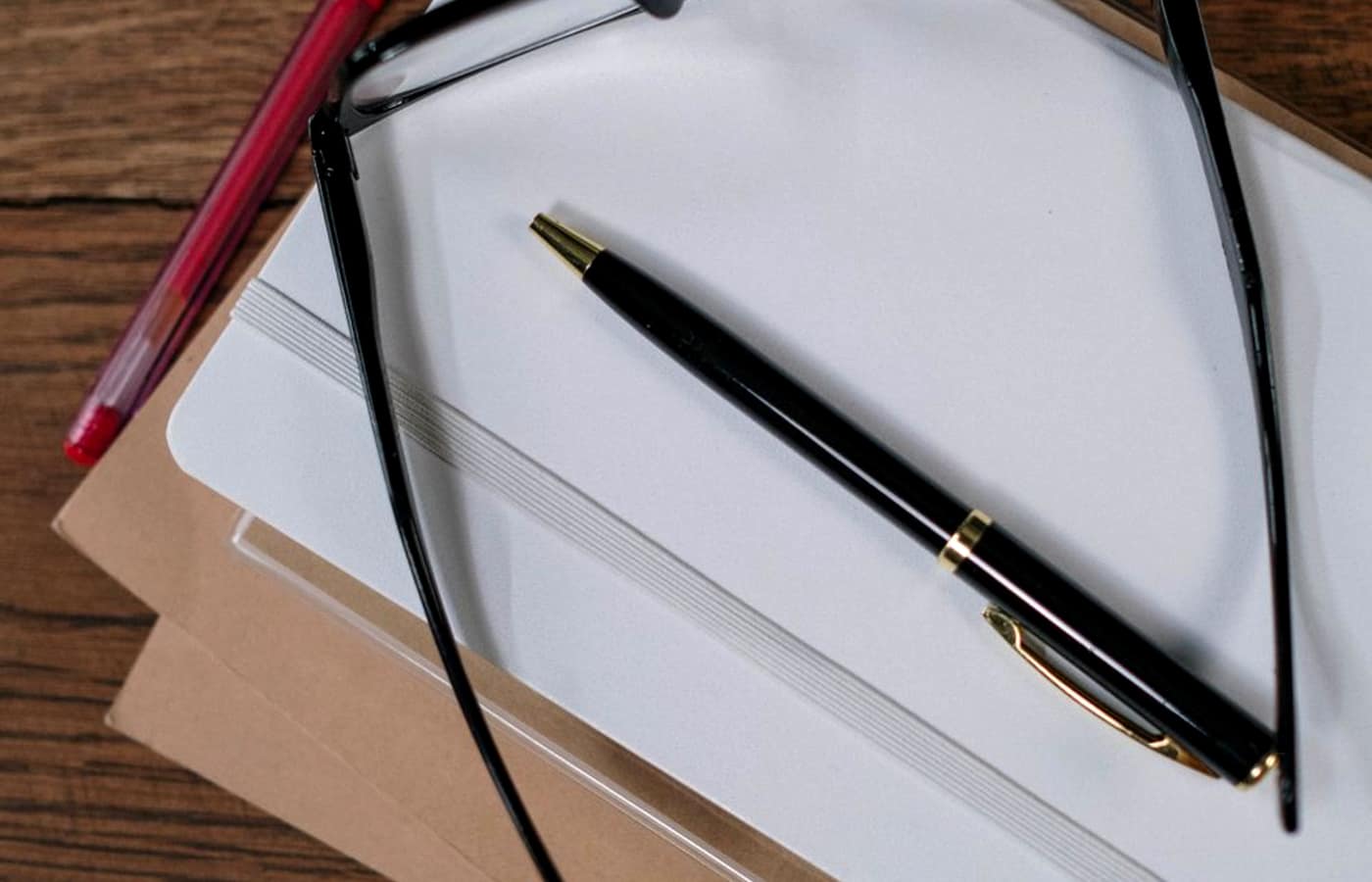 A pen and reading glasses resting on a blank notepad on a wooden desk.